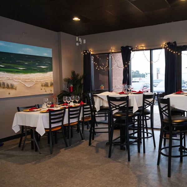 Restaurant dining room with set tables, a beach painting, and string lights by the window.