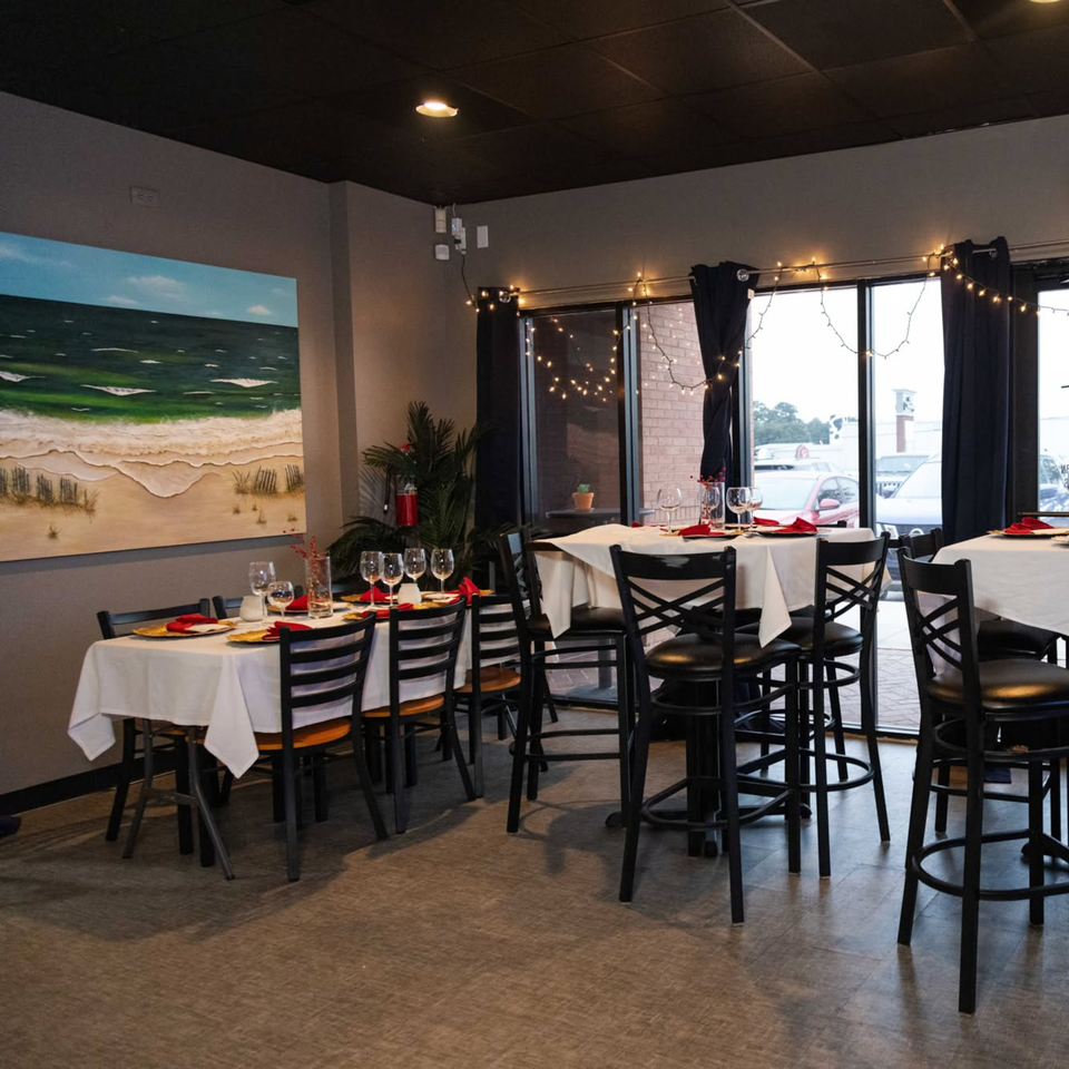 Restaurant dining room with set tables, a beach painting, and string lights by the window.