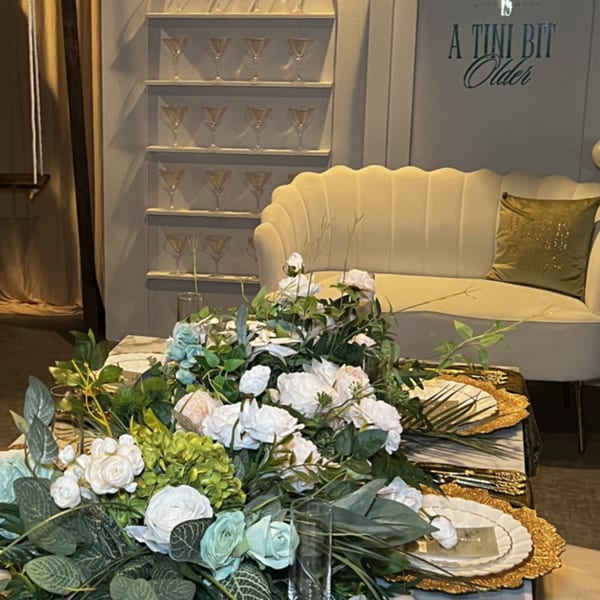 A table set with white and green floral arrangements, golden chargers, and a cream sofa in the background.