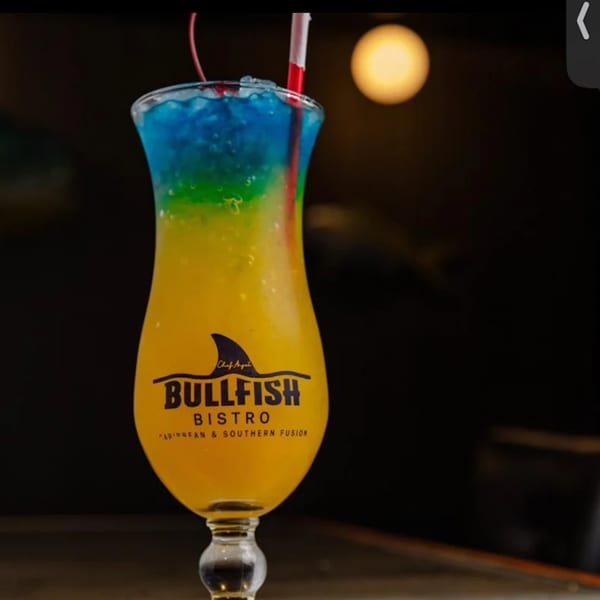 A Bullfish Bistro cocktail, blue, green, and yellow, with a red straw and cherry.