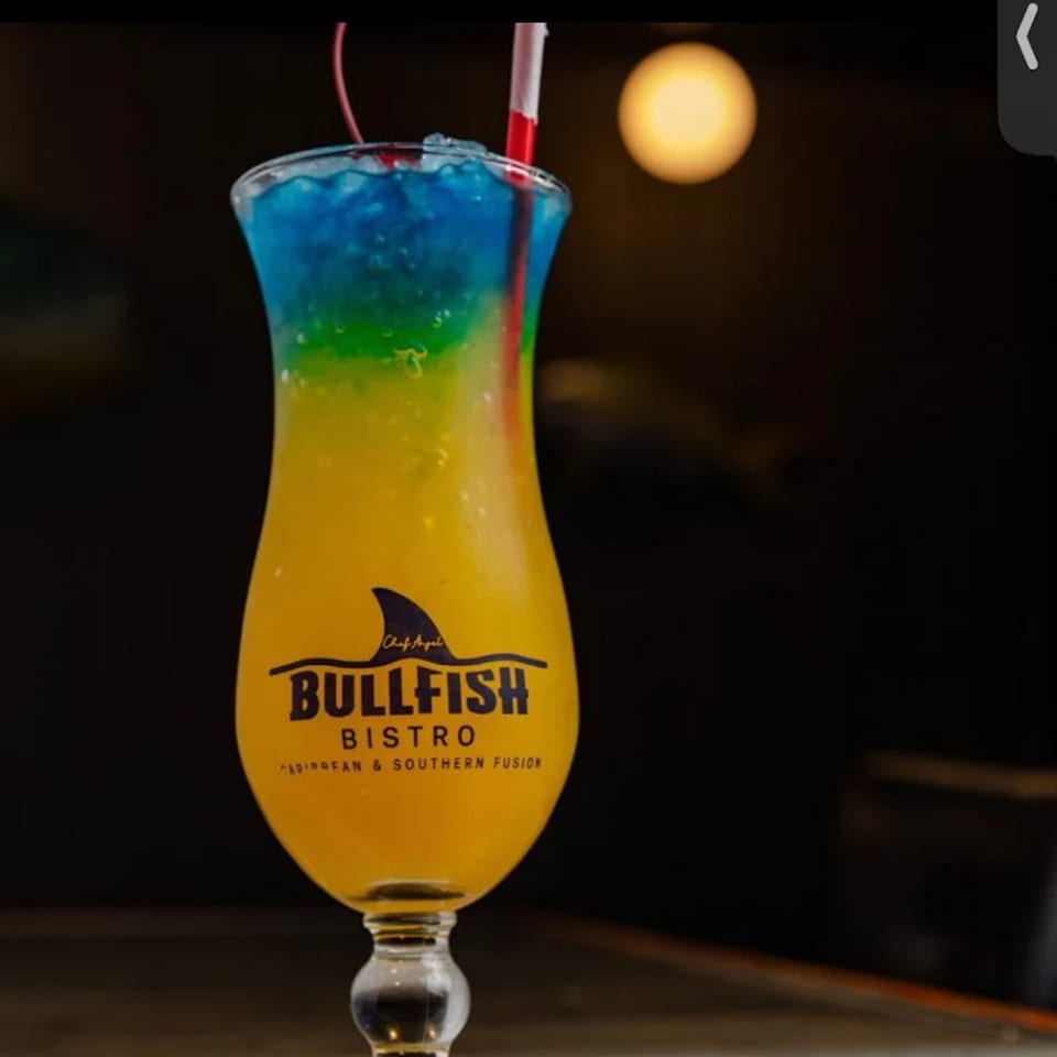 A Bullfish Bistro cocktail, blue, green, and yellow, with a red straw and cherry.