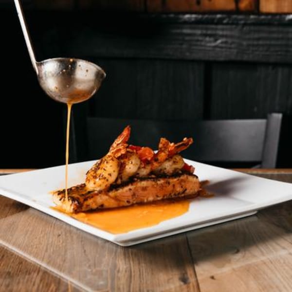 A ladle pours sauce over a Caribbean-style salmon and shrimp dish on a white plate.