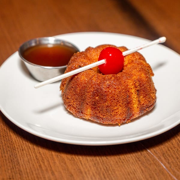 A golden-brown Caribbean rum cake with a red cherry on a white plate, next to a small cup of sauce.
